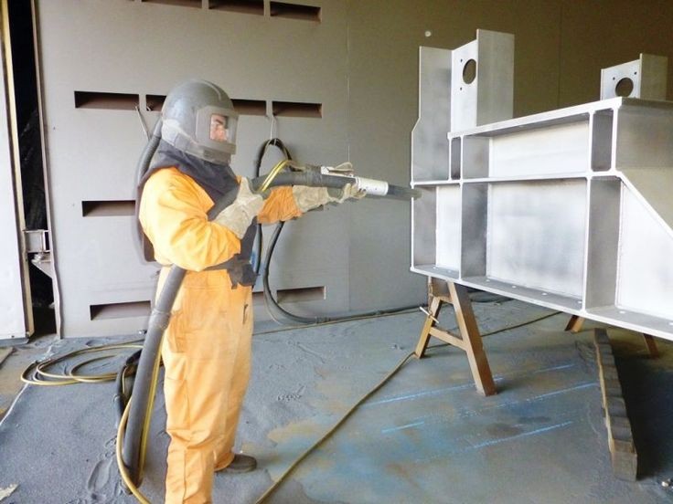 Opérateur en combinaison intégrale effectuant le sablage d'une structure métallique dans une cabine industrielle.